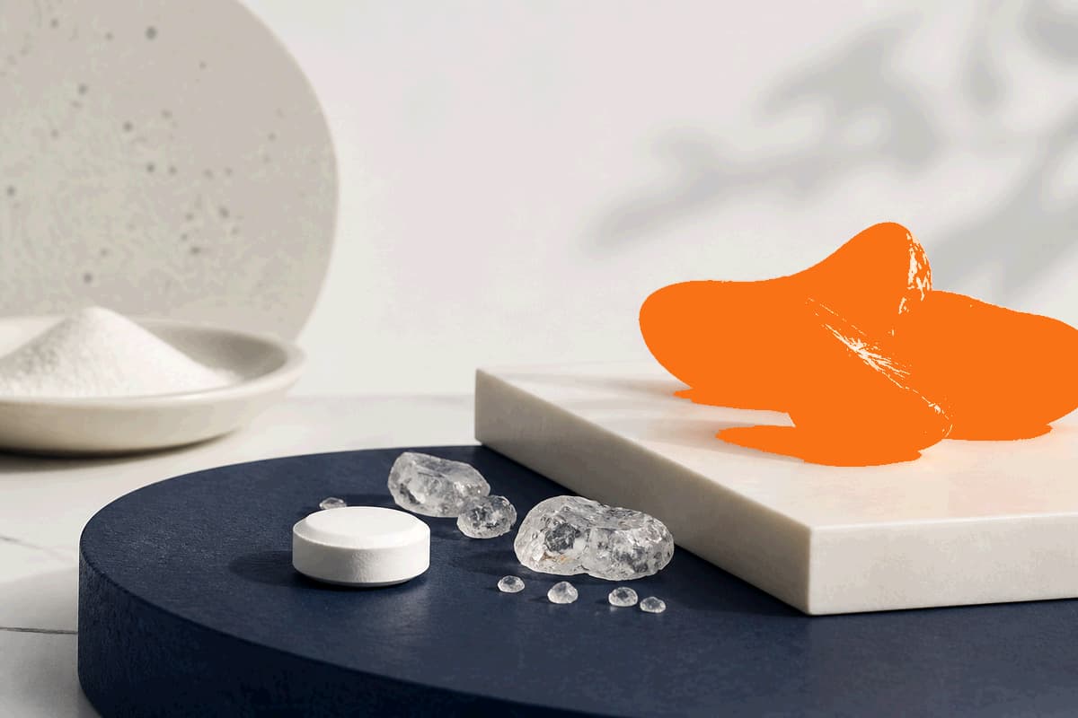 White vitamin C tablet, clear ascorbic acid crystals and stylized bright orange citrus segments on cream and deep navy surfaces